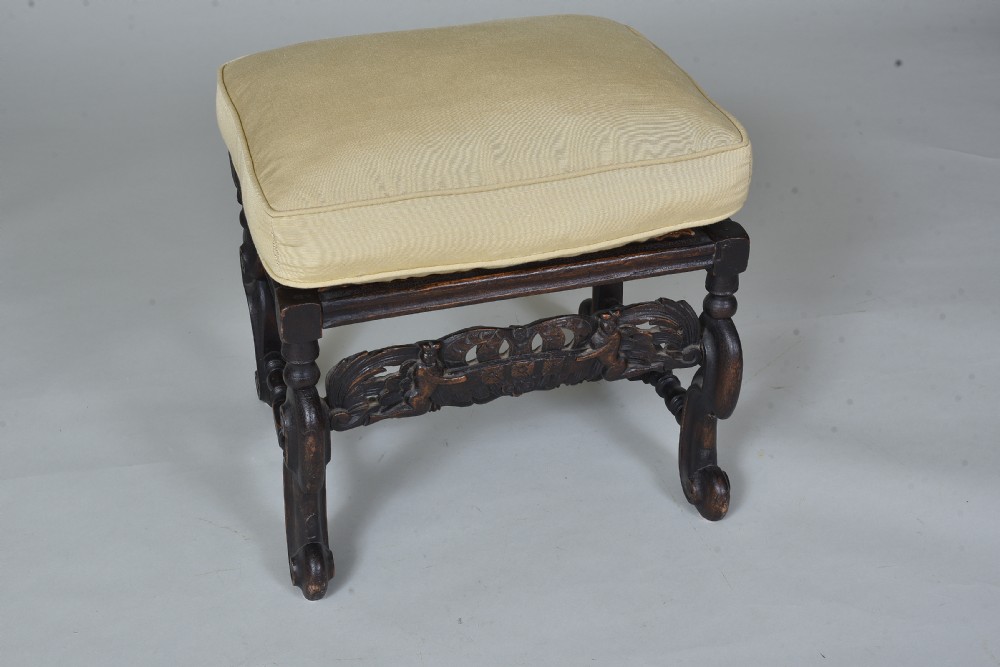 An antique dark brown carved wooden stool featuring scroll legs, an ornate carved apron, and a cream upholstered cushion. View III