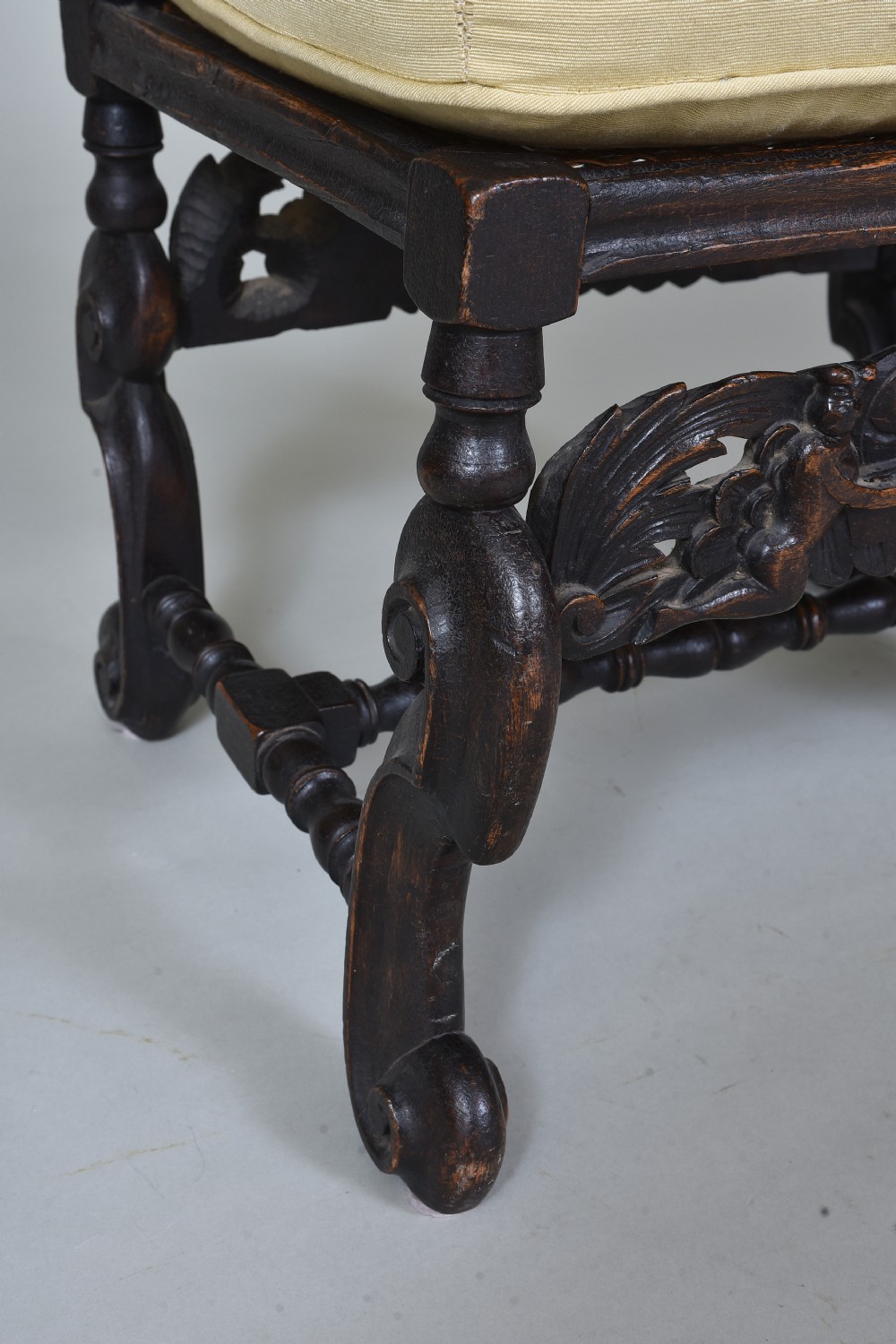 An antique dark brown carved wooden stool featuring scroll legs, an ornate carved apron, and a cream upholstered cushion. View IV
