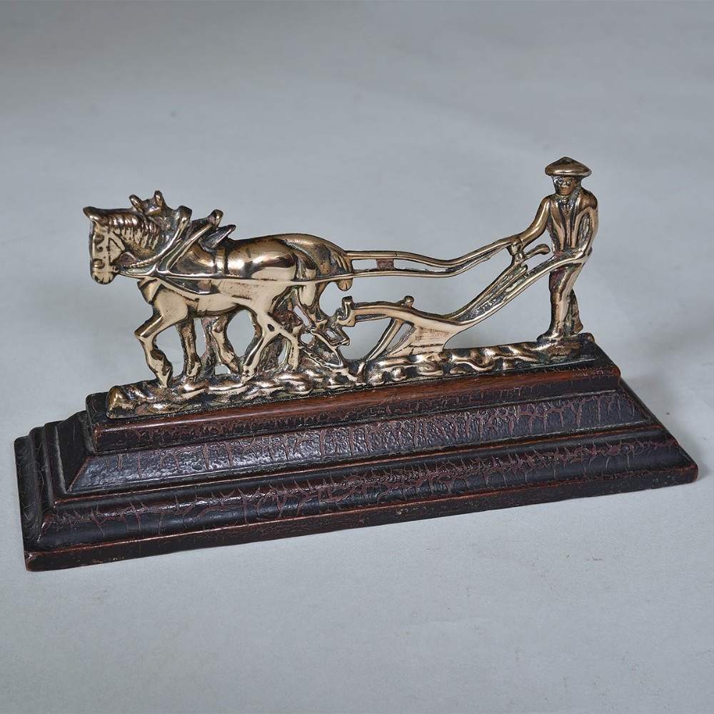 A cast brass figurine depicting a horse-drawn plough and farmer mounted on a dark textured wooden base.