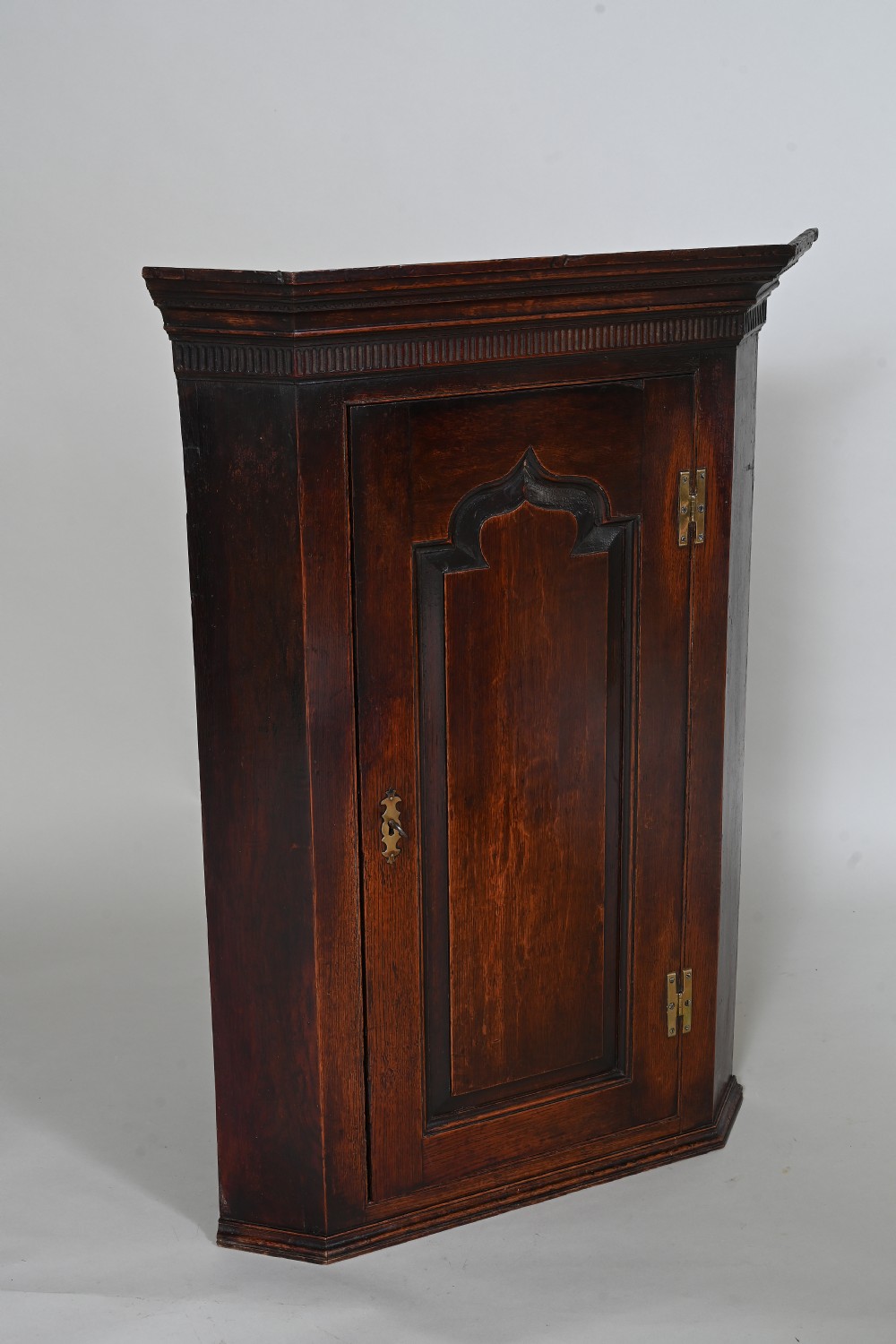 An eighteenth-century dark oak wall-hanging corner cupboard featuring an ogee-arched fielded panel door, brass H-hinges, brass keyhole escutcheon, and a moulded cornice.