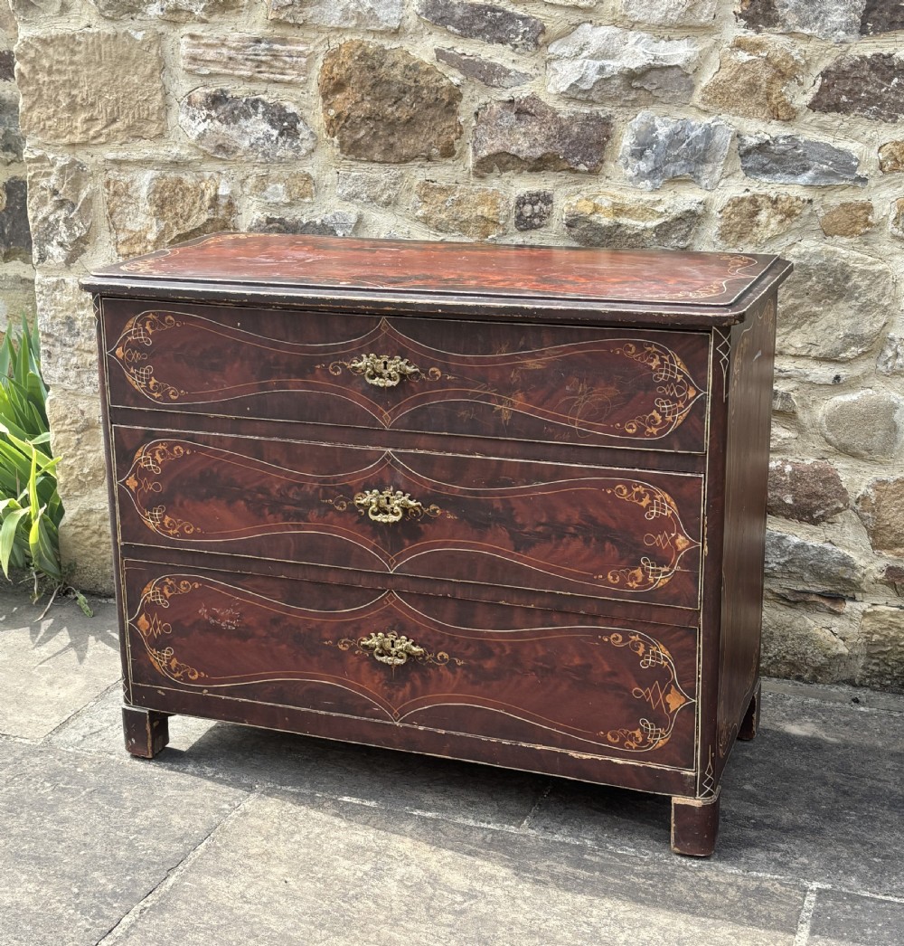 A three-drawer dark red painted wooden chest featuring gold filigree scrollwork, faux grain detailing, and ornate brass drop handles. View IV