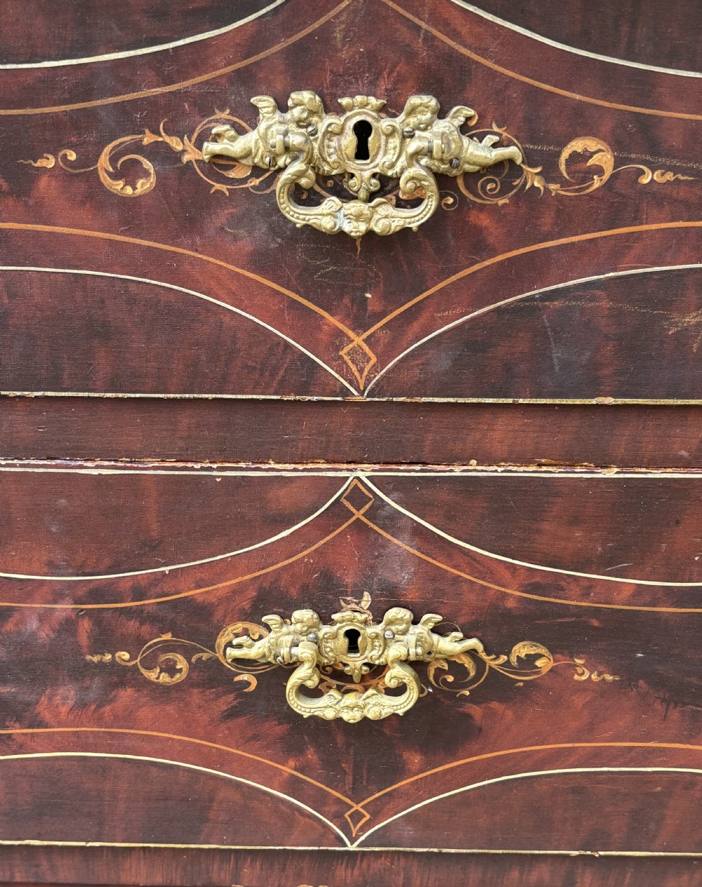 A three-drawer dark red painted wooden chest featuring gold filigree scrollwork, faux grain detailing, and ornate brass drop handles. View V