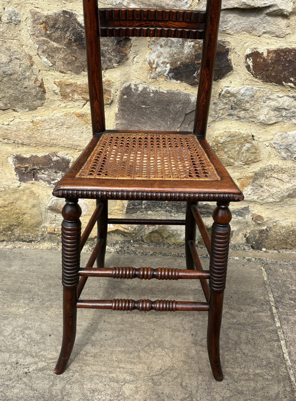 A tall dark wood childs high chair with a woven cane seat, reeded back slats, and bobbin turned front legs. View II
