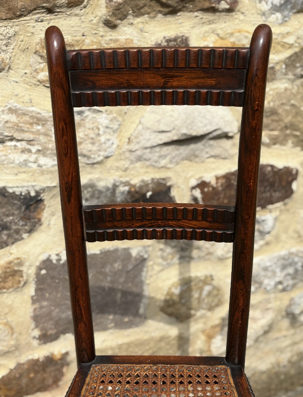 A tall dark wood childs high chair with a woven cane seat, reeded back slats, and bobbin turned front legs. View III