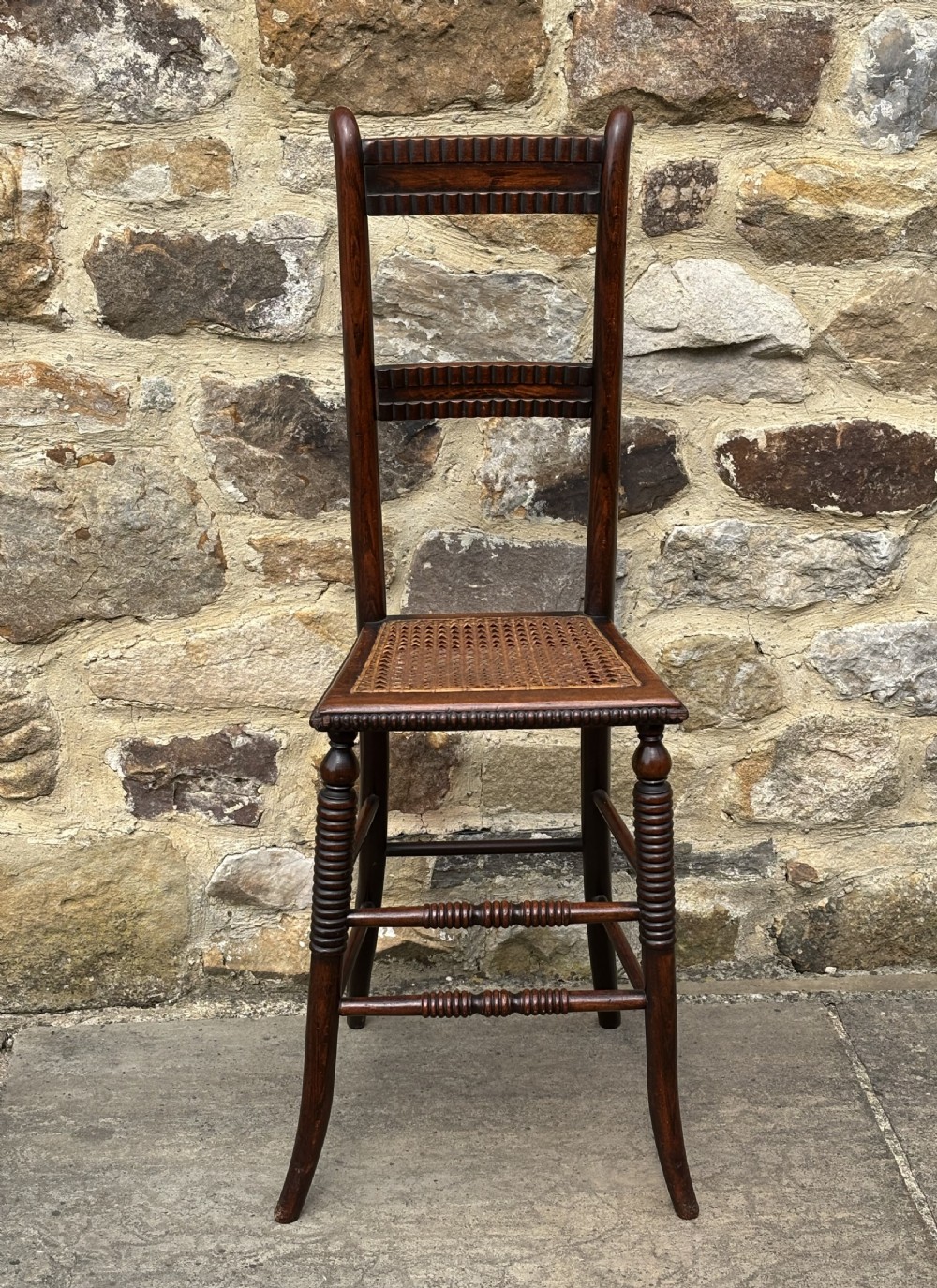 A tall dark wood childs high chair with a woven cane seat, reeded back slats, and bobbin turned front legs. View IV
