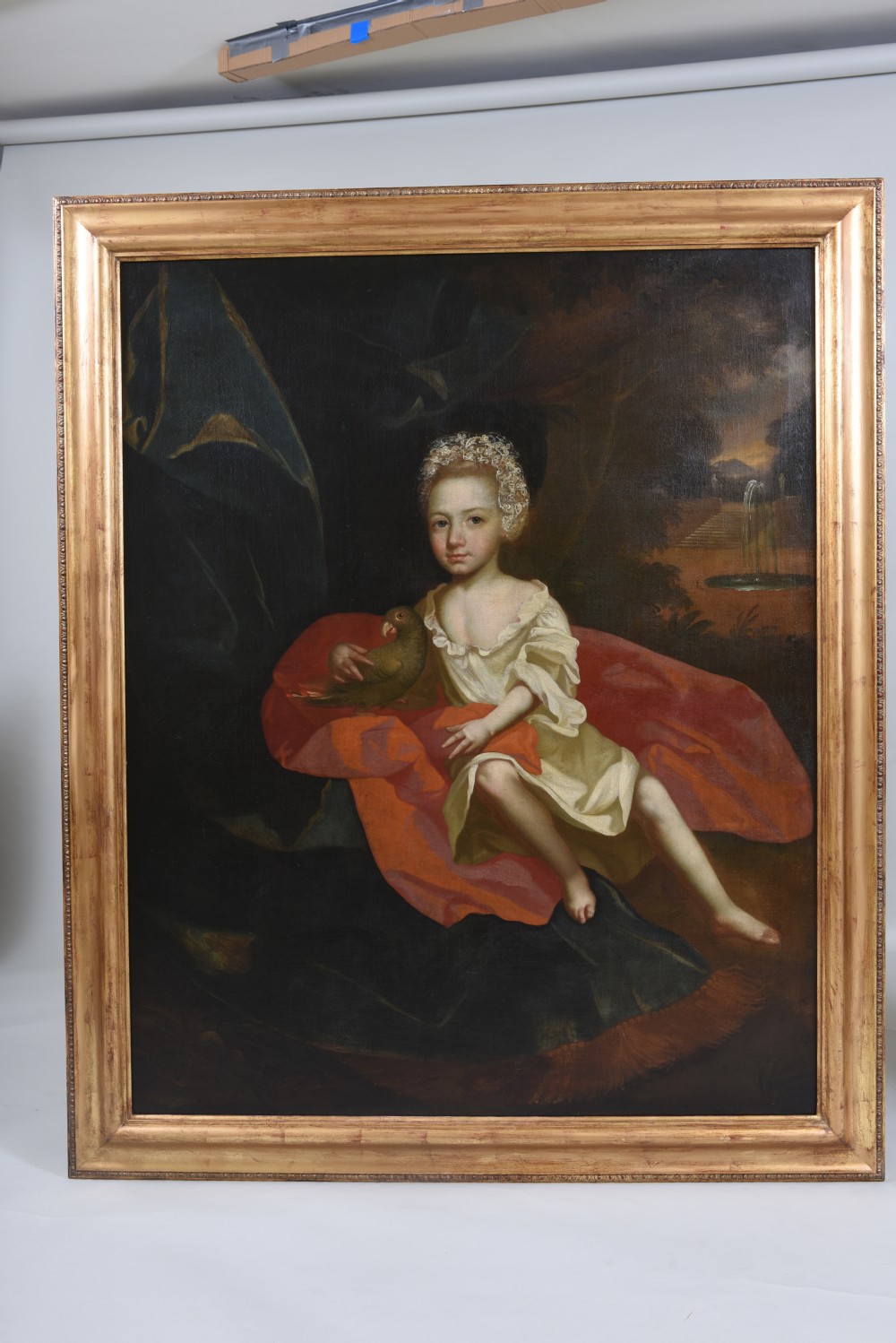 An 18th-century oil portrait depicting a young girl in a white dress seated beside a red drapery holding a bird, enclosed in a giltwood frame. View II
