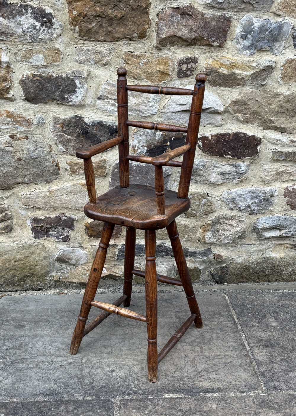 An antique dark timber folk art child high chair featuring turned legs, a footrest, and armrests against a stone wall.