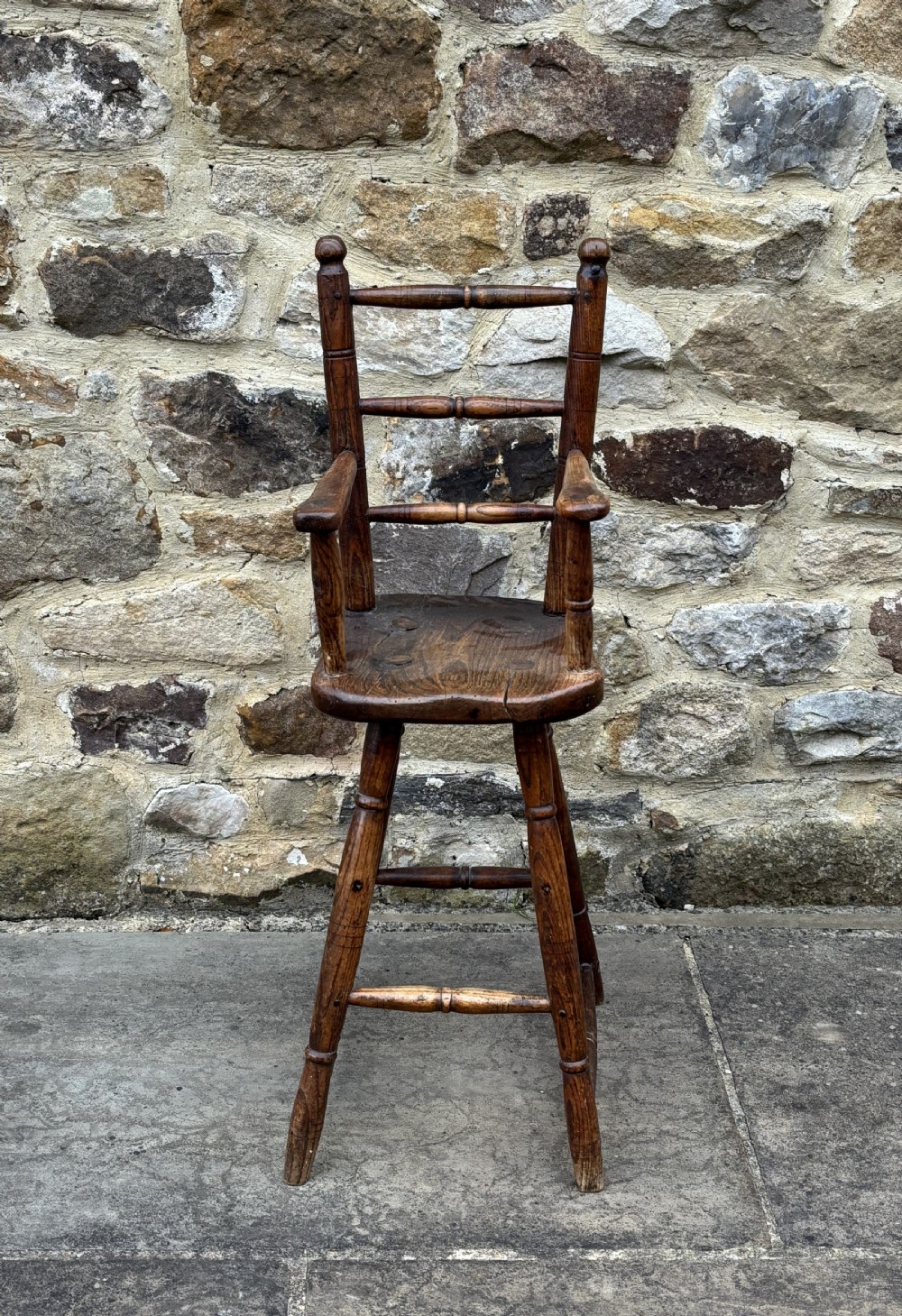 An antique dark timber folk art child high chair featuring turned legs, a footrest, and armrests against a stone wall. View II