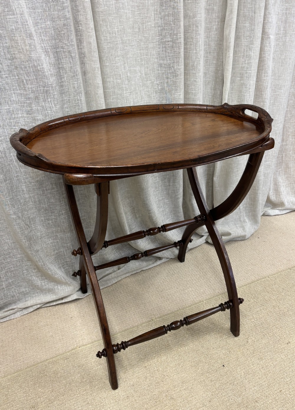 An antique dark oak oval butlers tray with carved edge detailing resting on a matching folding stand with curved legs and turned stretchers.