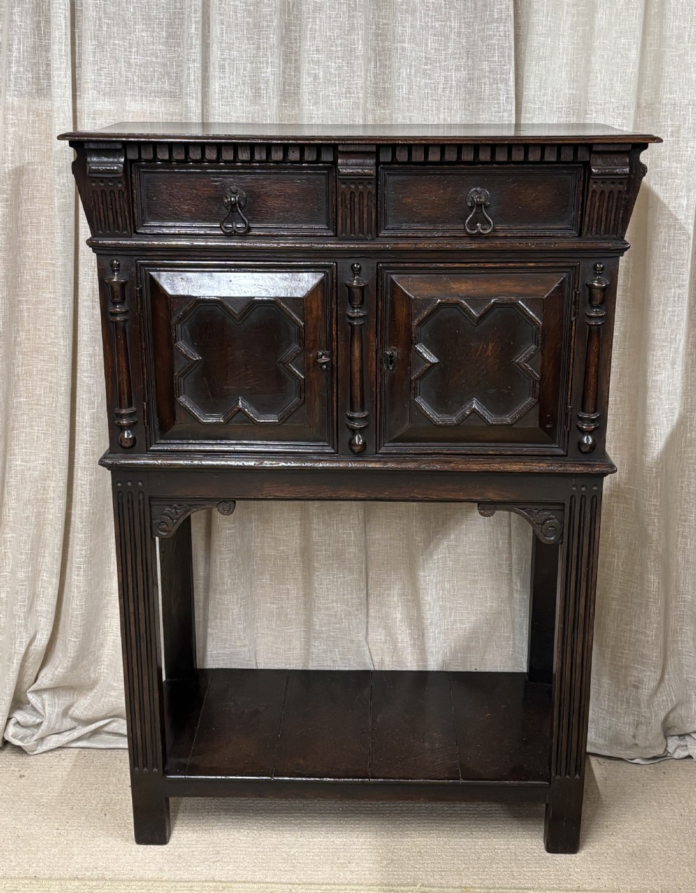 A dark carved oak cupboard raised on an open stand with two top drawers, geometric moulded doors, and a bottom shelf.