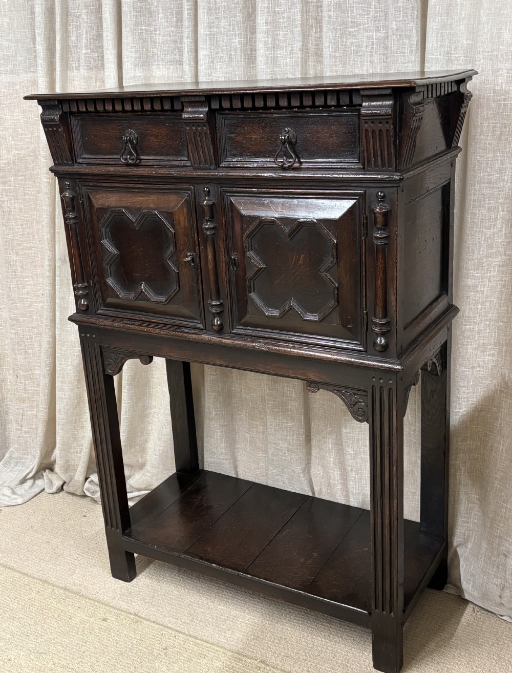 A dark carved oak cupboard raised on an open stand with two top drawers, geometric moulded doors, and a bottom shelf. View II
