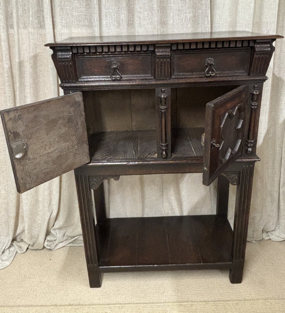 A dark carved oak cupboard raised on an open stand with two top drawers, geometric moulded doors, and a bottom shelf. View III
