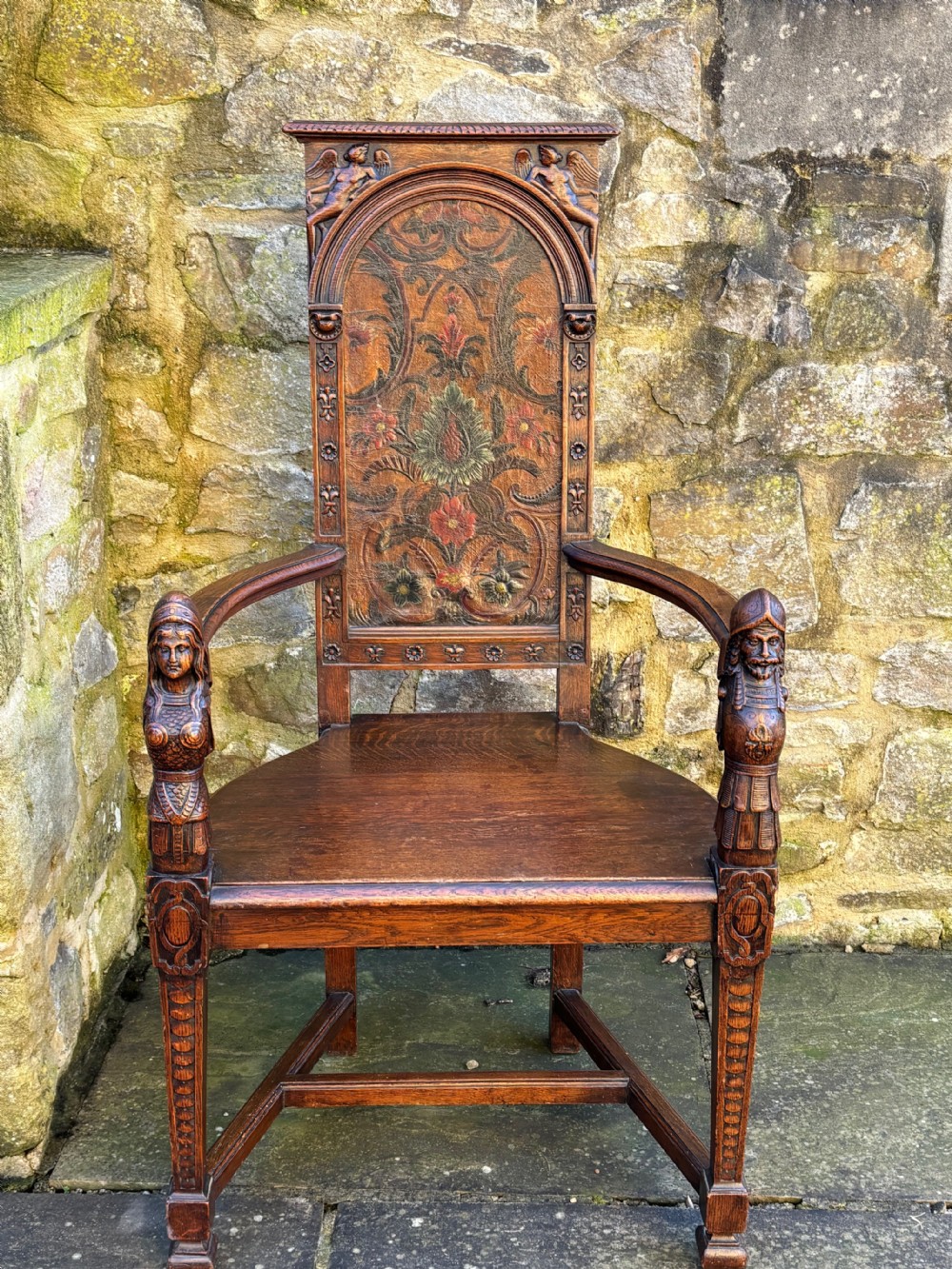 A 19th-century carved dark oak caquetoire armchair featuring a trapezoidal seat, carved armrest figures, and an arched polychrome back panel.