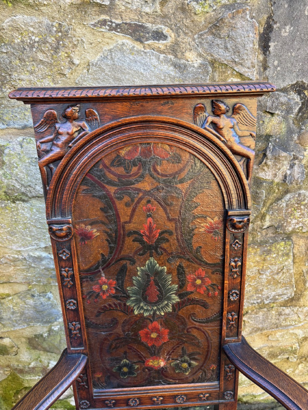A 19th-century carved dark oak caquetoire armchair featuring a trapezoidal seat, carved armrest figures, and an arched polychrome back panel. View V