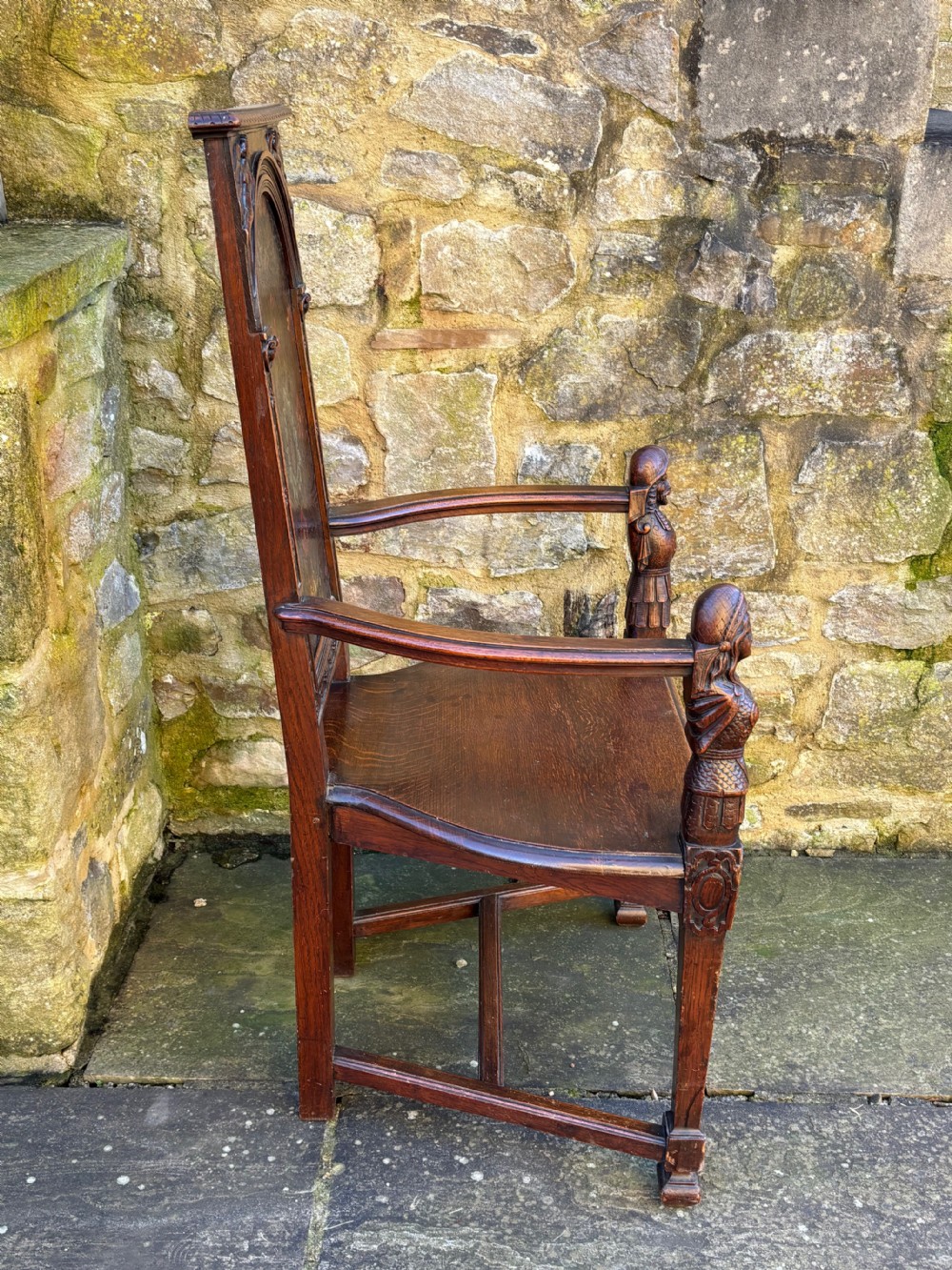 A 19th-century carved dark oak caquetoire armchair featuring a trapezoidal seat, carved armrest figures, and an arched polychrome back panel. View VI