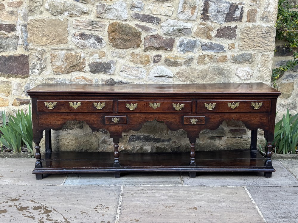 An eighteenth-century dark oak potboard dresser base featuring brass batwing handles, a shaped apron, turned front legs, and an open lower shelf.