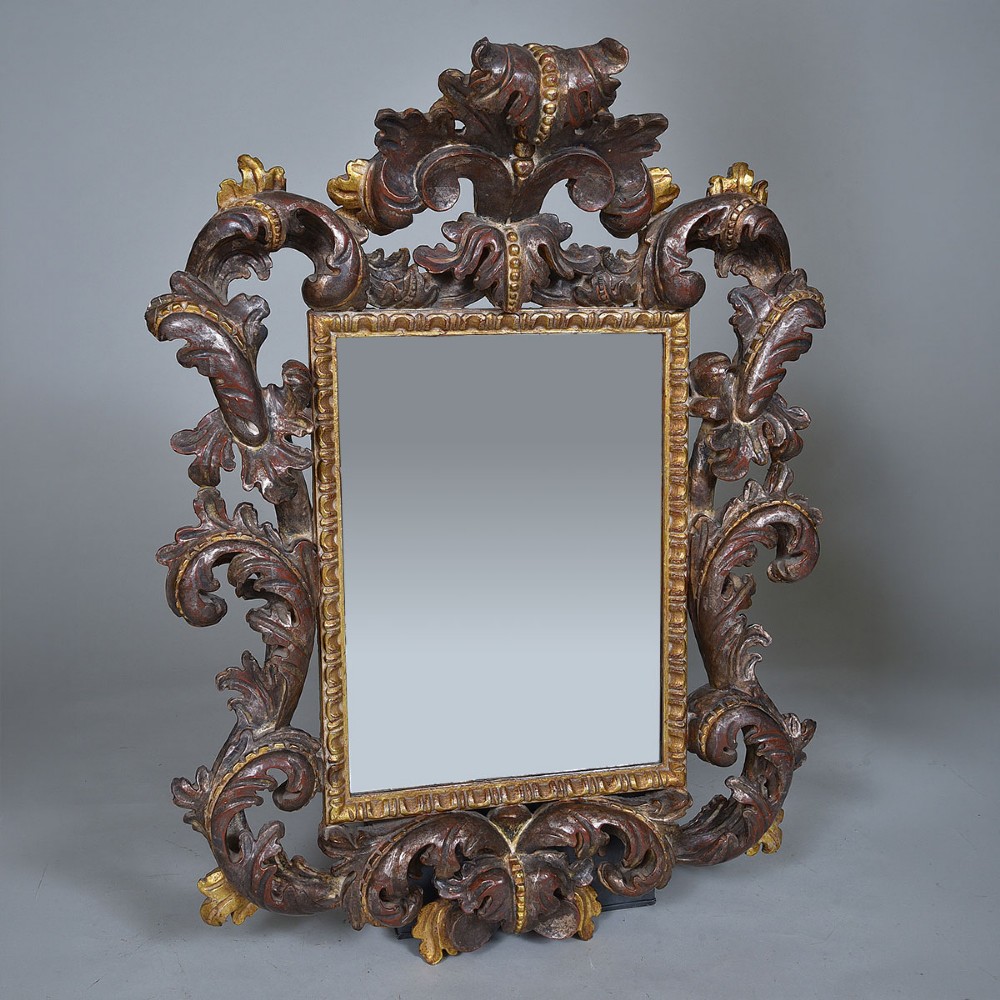 An ornate Italian carved wood wall mirror with elaborate acanthus leaf cresting and C-scroll motifs finished in gold and silver leafing around a central glass plate.
