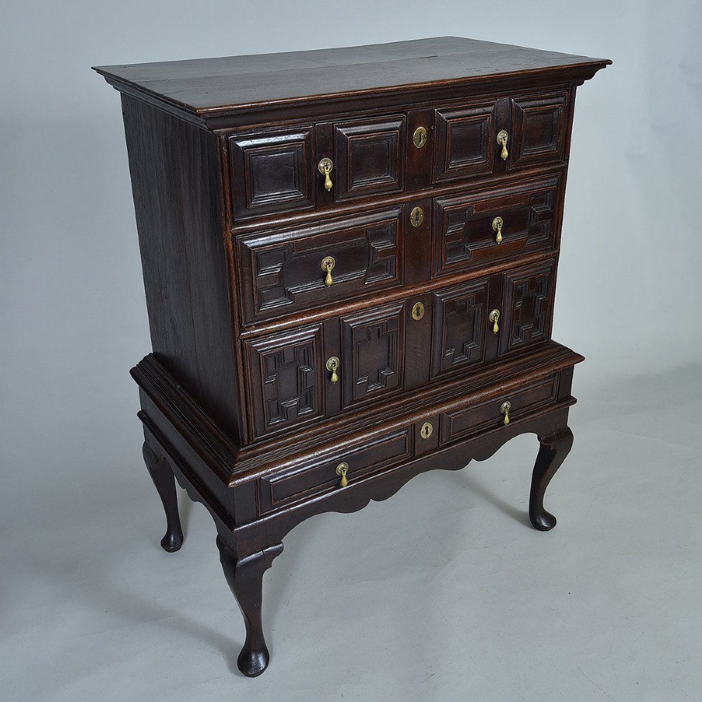 A dark oak chest on stand with geometric panelled drawers, brass drop handles, and cabriole legs. View VII