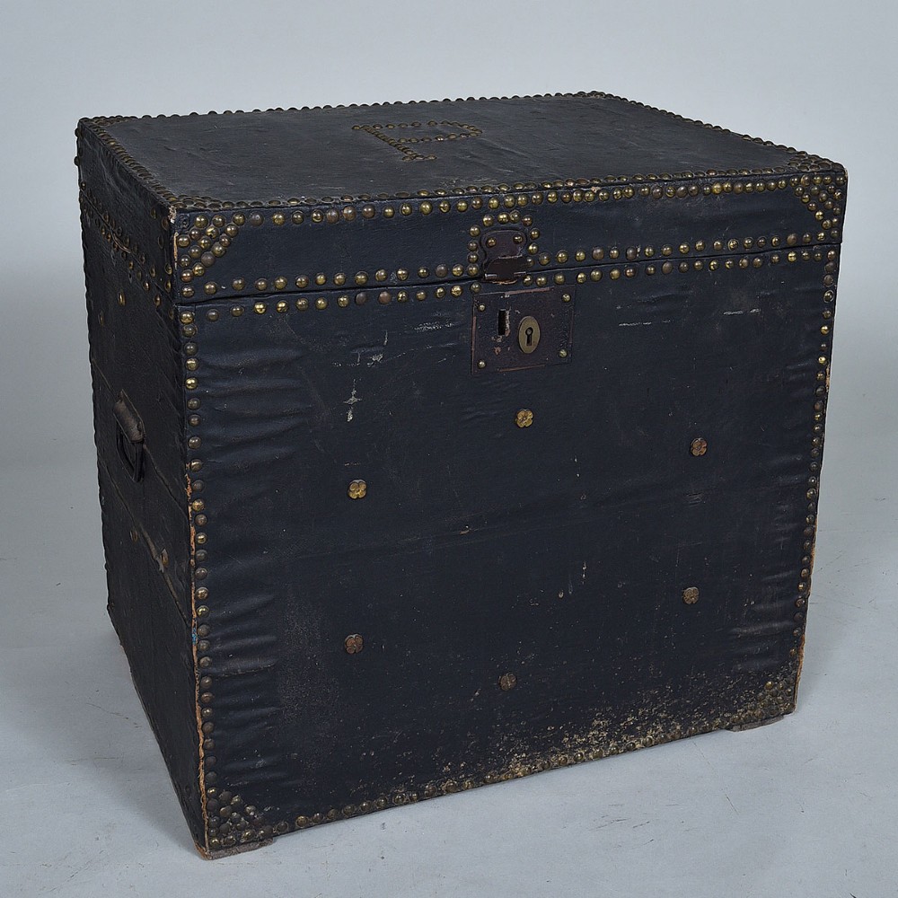 A dark leather-covered antique trunk featuring dense brass stud embellishments, a monogrammed lid, and a central brass lock plate.