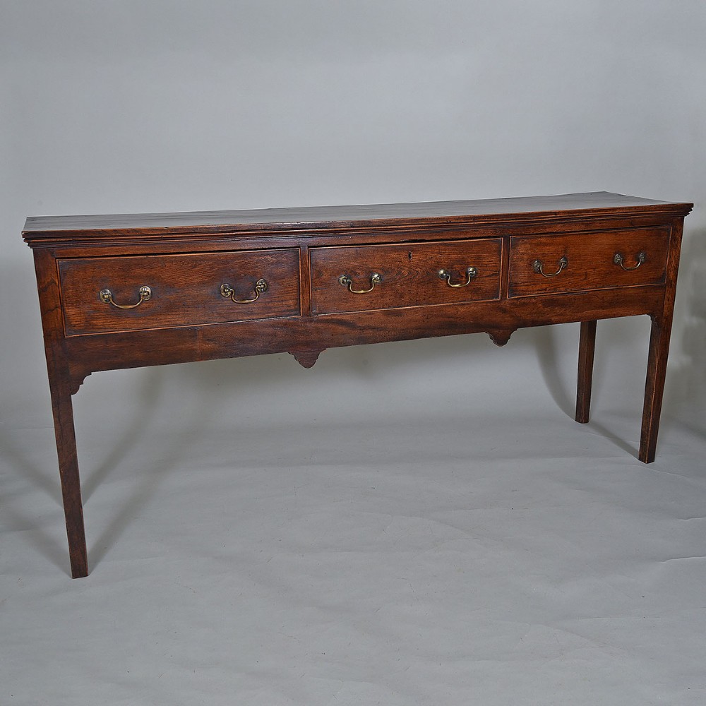 An 18th-century warm brown elm dresser base featuring three drawers with brass drop handles and square legs.