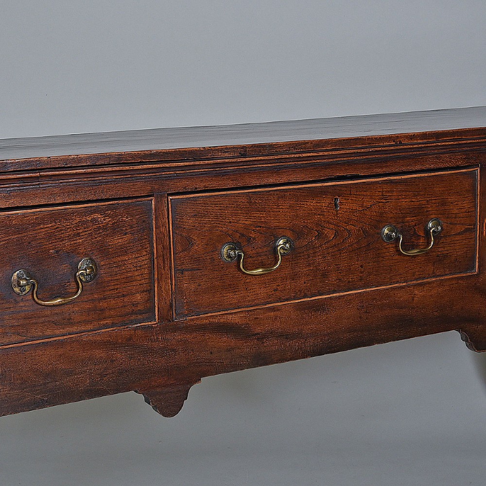 An 18th-century warm brown elm dresser base featuring three drawers with brass drop handles and square legs. View II