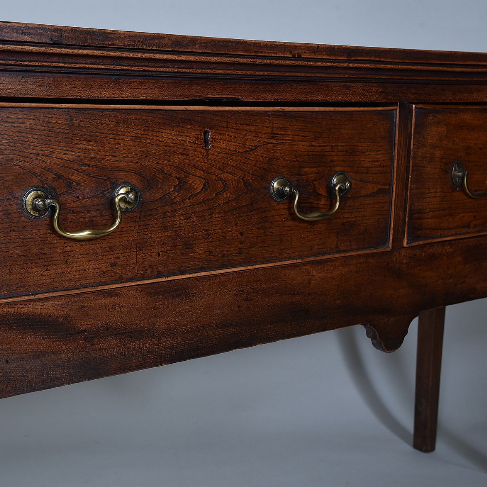 An 18th-century warm brown elm dresser base featuring three drawers with brass drop handles and square legs. View IV