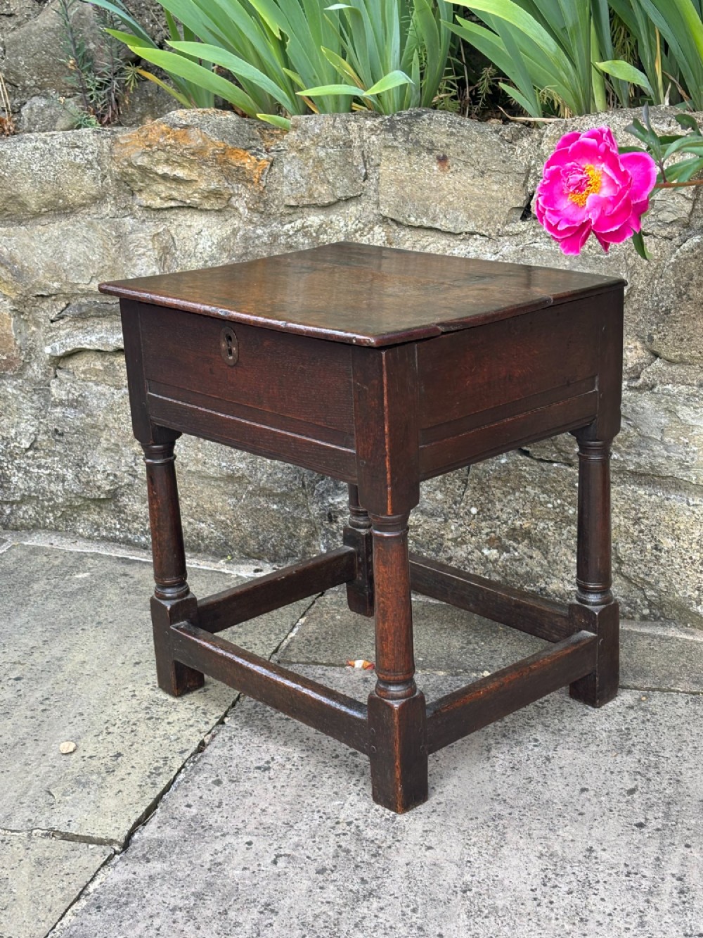 A dark oak 17th-century box stool featuring a flat hinged rectangular lid, a keyhole on the front apron, turned legs, and lower wooden stretchers. View II