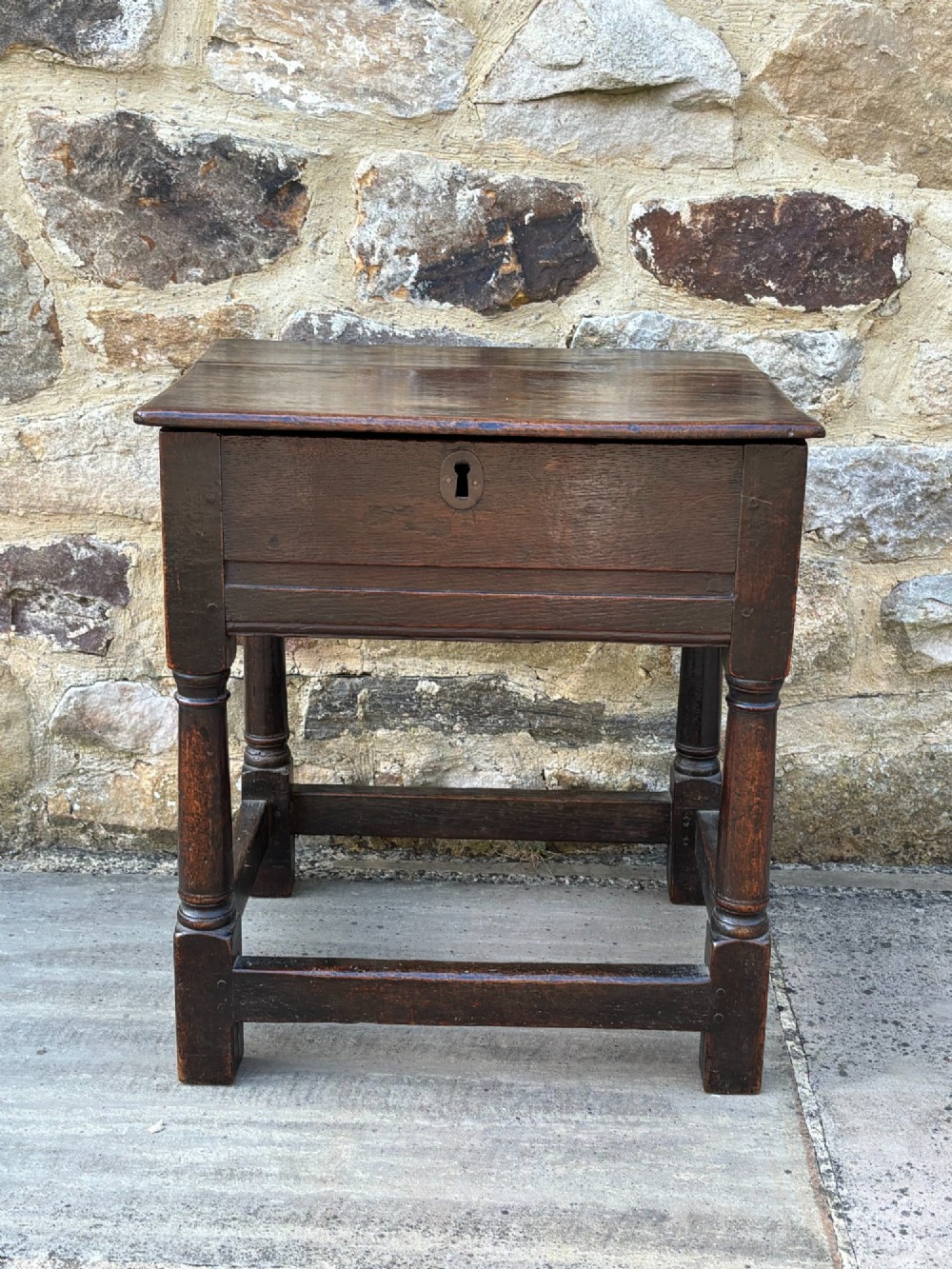 A dark oak 17th-century box stool featuring a flat hinged rectangular lid, a keyhole on the front apron, turned legs, and lower wooden stretchers. View III