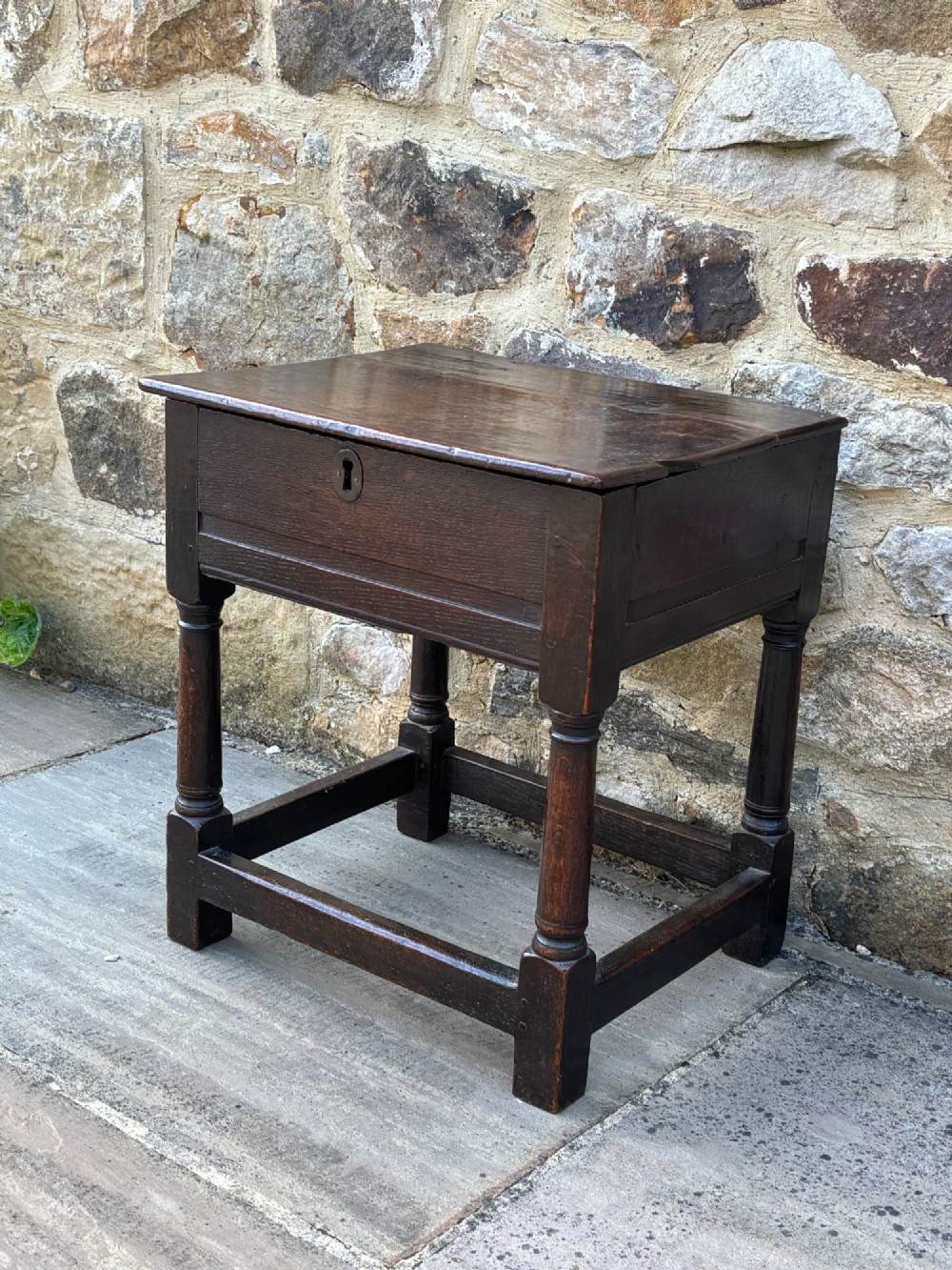 A dark oak 17th-century box stool featuring a flat hinged rectangular lid, a keyhole on the front apron, turned legs, and lower wooden stretchers. View IV