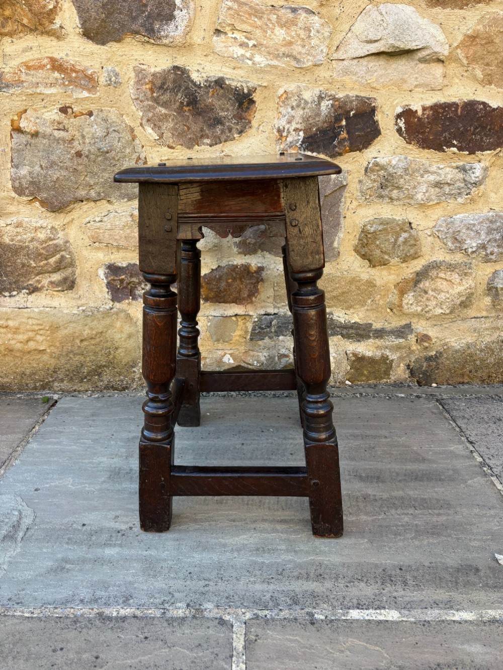 A 17th-century dark oak joint stool featuring turned legs, pegged mortise and tenon joinery, a carved frieze, and a plain rectangular wooden seat. View III