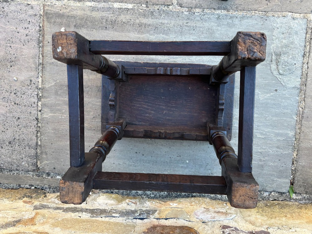 A 17th-century dark oak joint stool featuring turned legs, pegged mortise and tenon joinery, a carved frieze, and a plain rectangular wooden seat. View IV