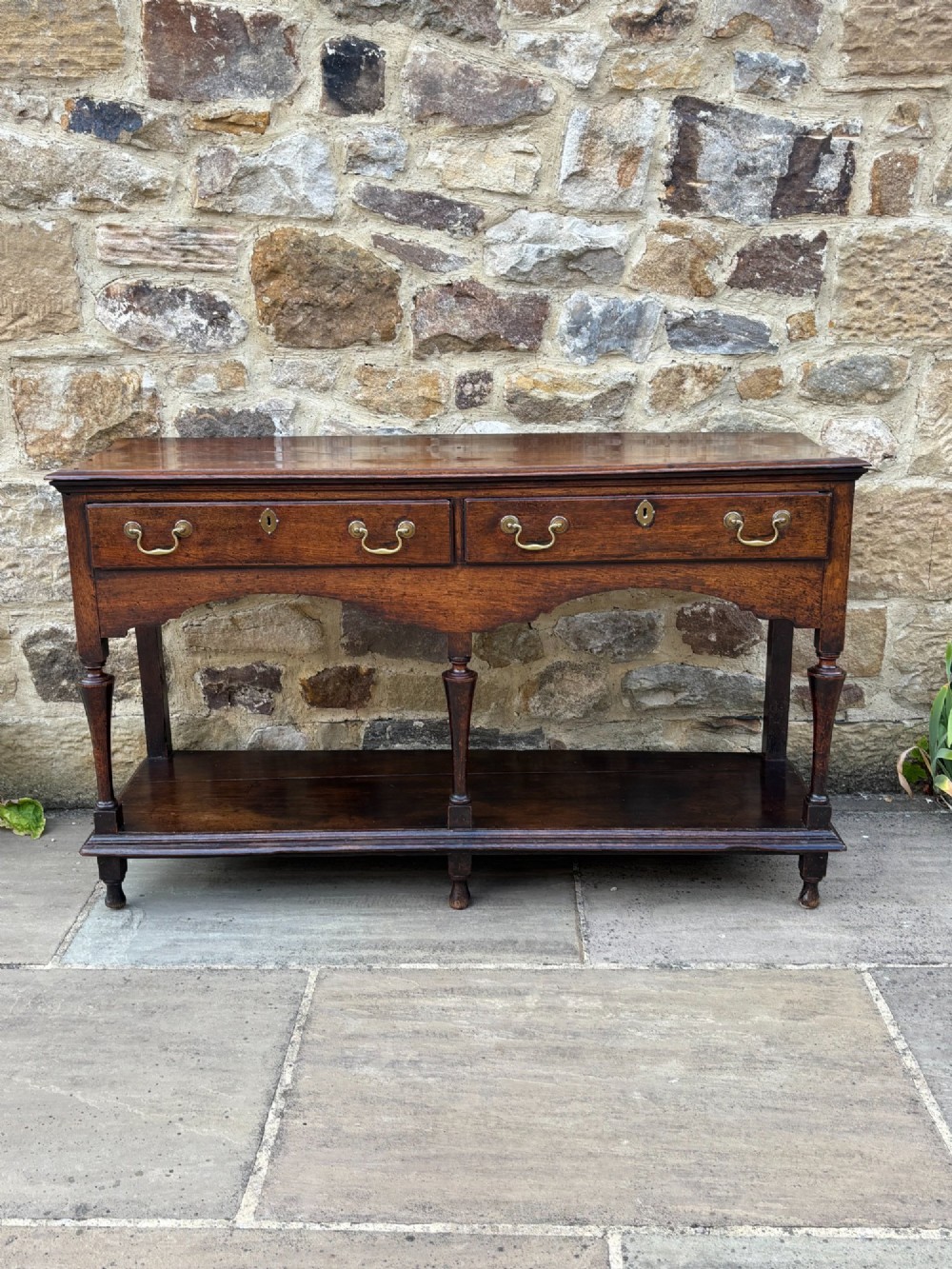 An 18th-century dark oak dresser base featuring two drawers with brass handles, a shaped apron, turned front legs, and a lower potboard shelf.