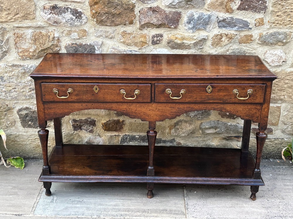 An 18th-century dark oak dresser base featuring two drawers with brass handles, a shaped apron, turned front legs, and a lower potboard shelf. View II