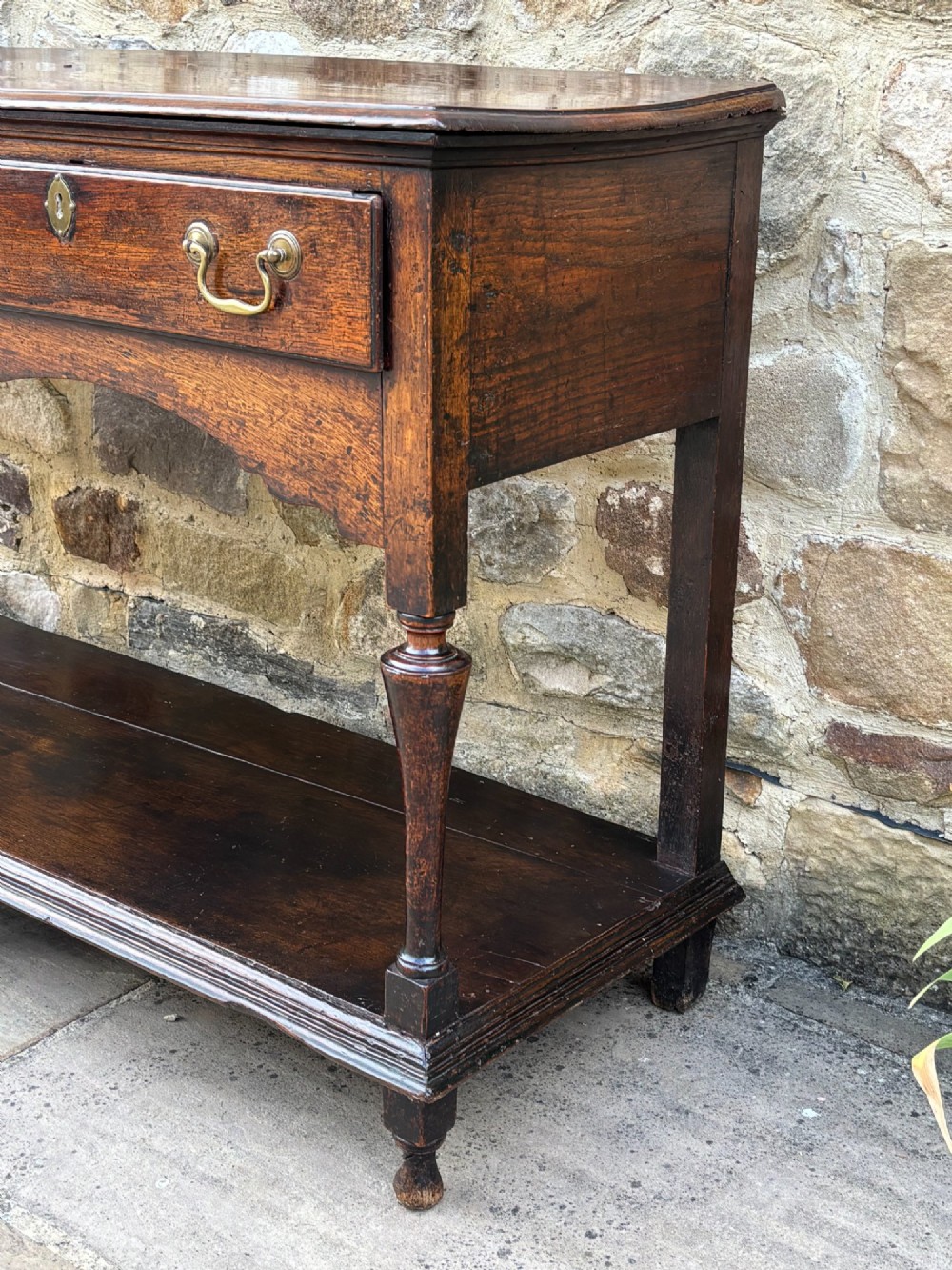An 18th-century dark oak dresser base featuring two drawers with brass handles, a shaped apron, turned front legs, and a lower potboard shelf. View III