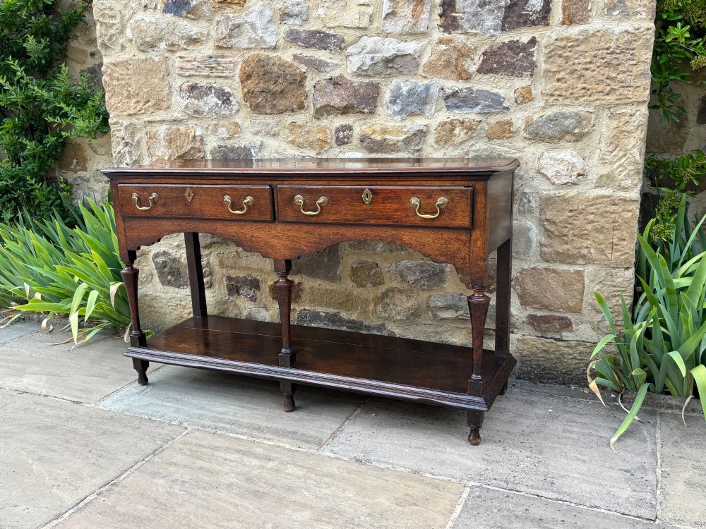 An 18th-century dark oak dresser base featuring two drawers with brass handles, a shaped apron, turned front legs, and a lower potboard shelf. View VI
