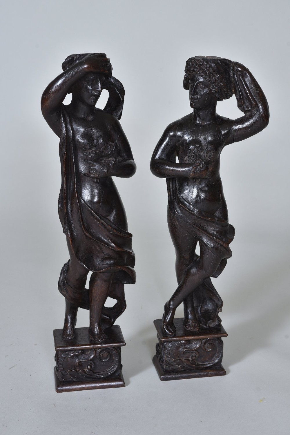 A pair of dark carved oak figures depicting classical drapery-clad subjects standing on carved square pedestals.