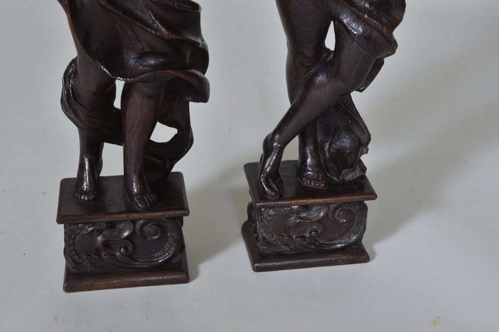 A pair of dark carved oak figures depicting classical drapery-clad subjects standing on carved square pedestals. View III