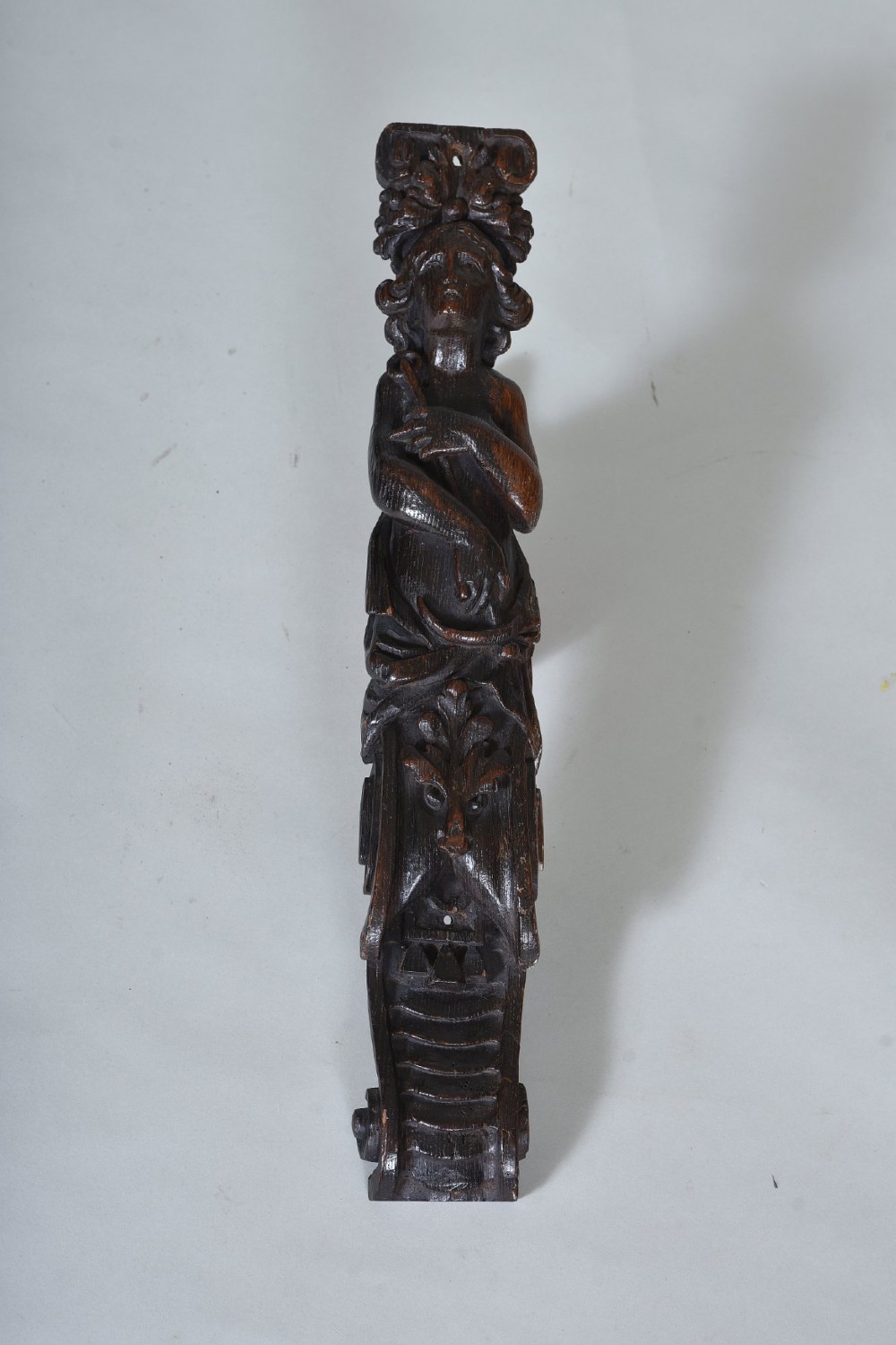 A dark oak 17th-century vertical wood carving featuring a female figure positioned above a carved grotesque mask.