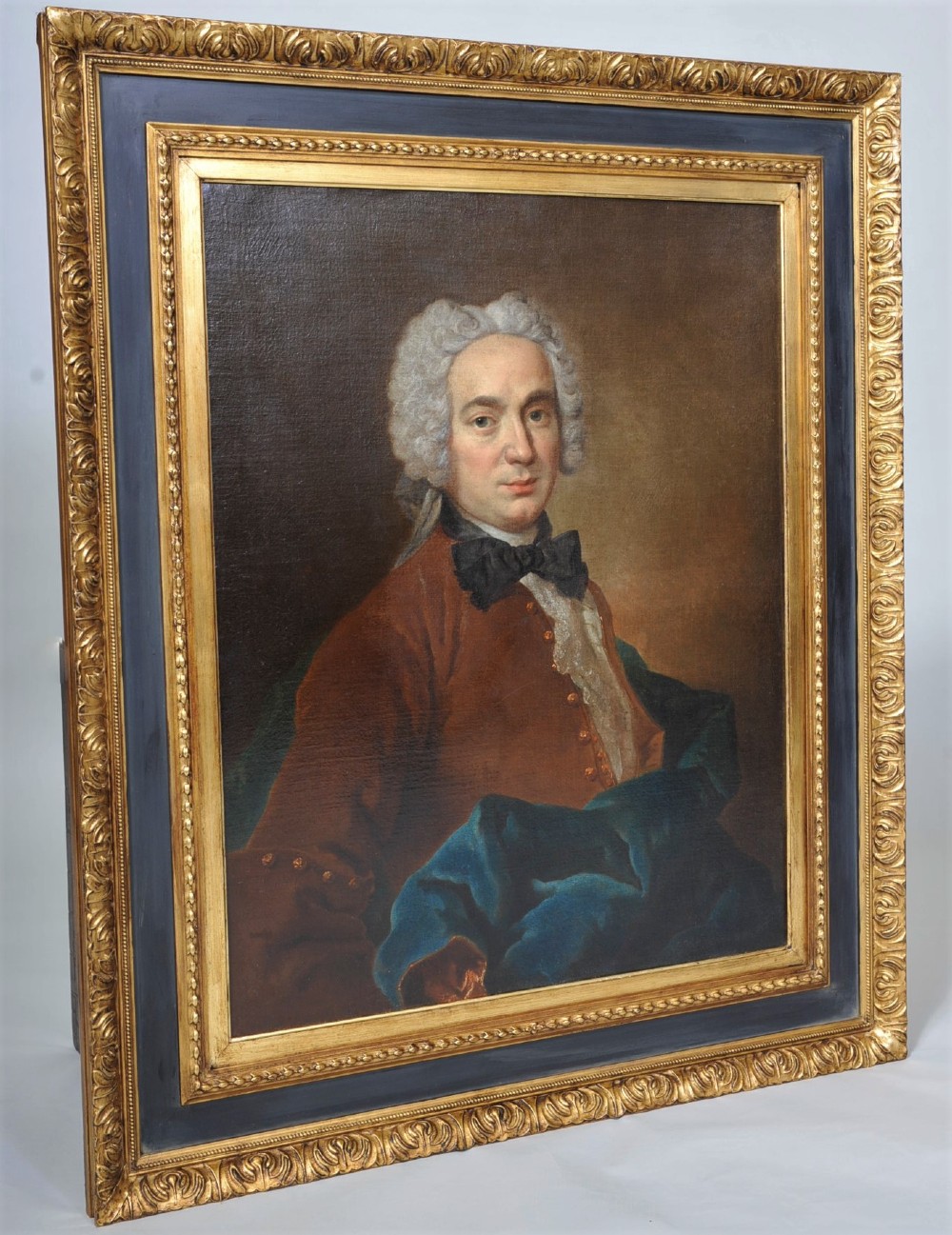 An eighteenth-century oil painting depicting a gentleman wearing a powdered wig, brown coat, black cravat, and blue drape, set within a carved gilt and dark wood frame.