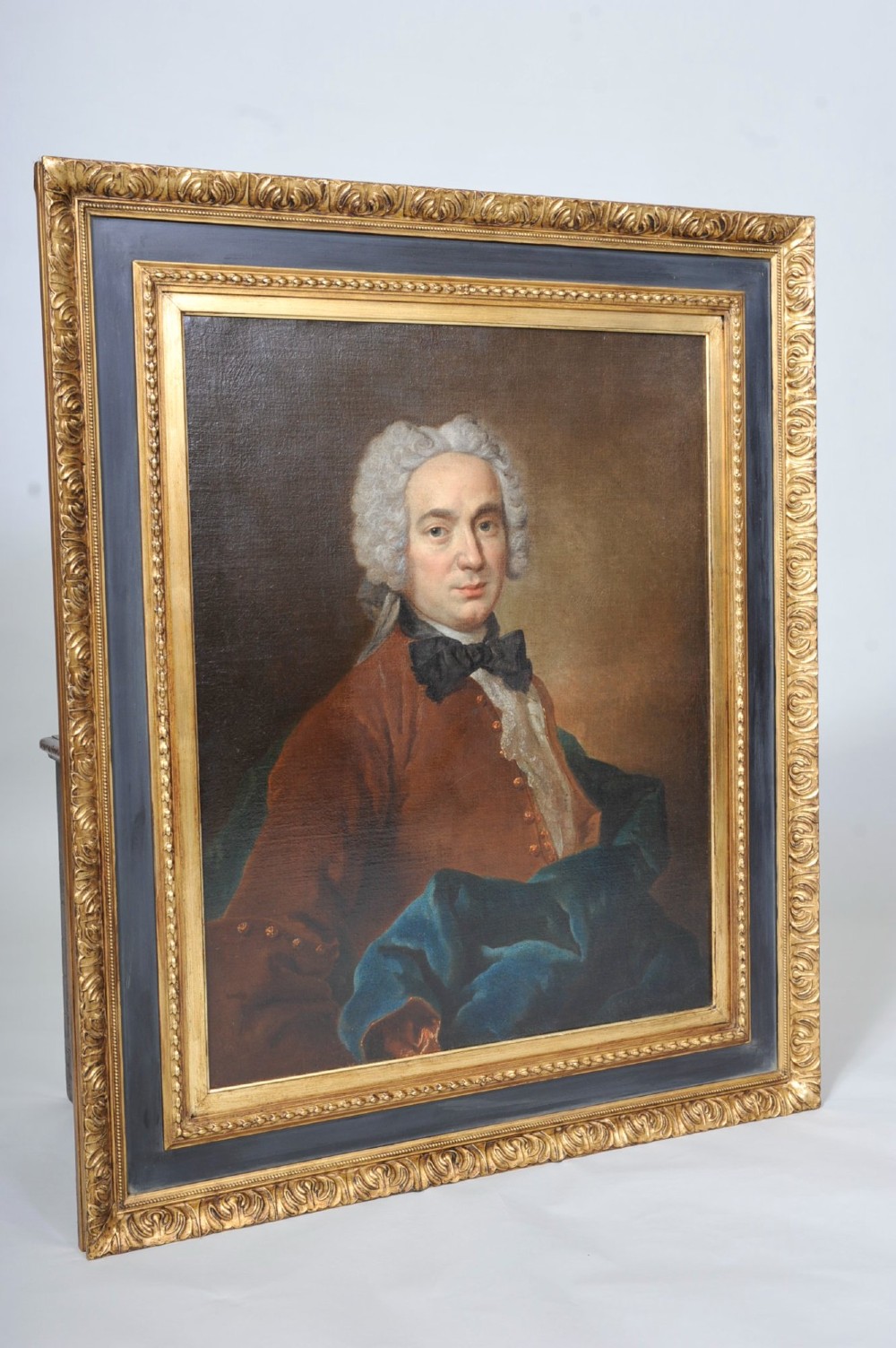 An eighteenth-century oil painting depicting a gentleman wearing a powdered wig, brown coat, black cravat, and blue drape, set within a carved gilt and dark wood frame. View II