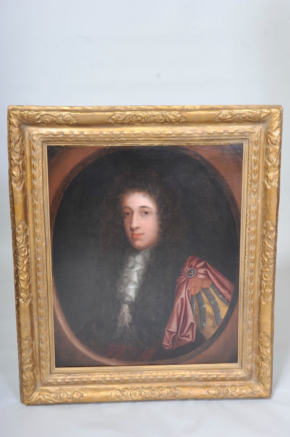 An oil painting depicting a 17th-century gentleman with a dark periwig and lace cravat set within an oval spandrel and a carved gilded frame.