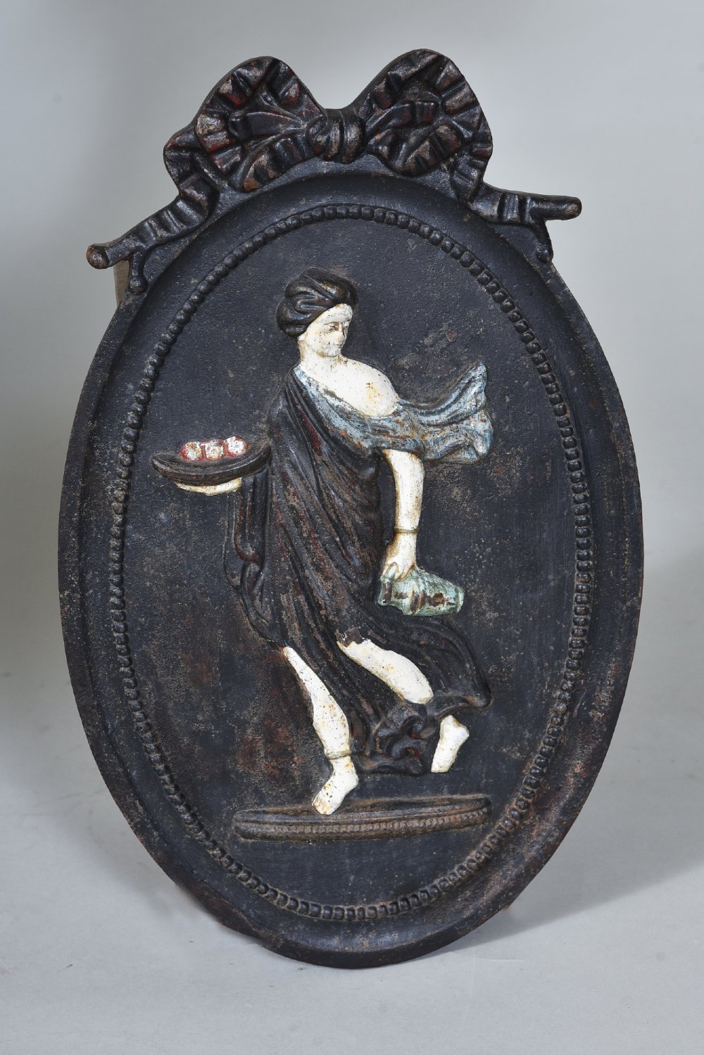 An oval dark cast iron wall plaque topped with a ribbon crest, depicting a classical female figure in draped robes painted in white and muted tones.