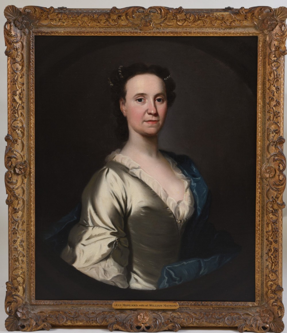An 18th-century oil painting on canvas depicting a seated woman in a cream satin dress with a blue wrap, set within an ornate carved giltwood frame.