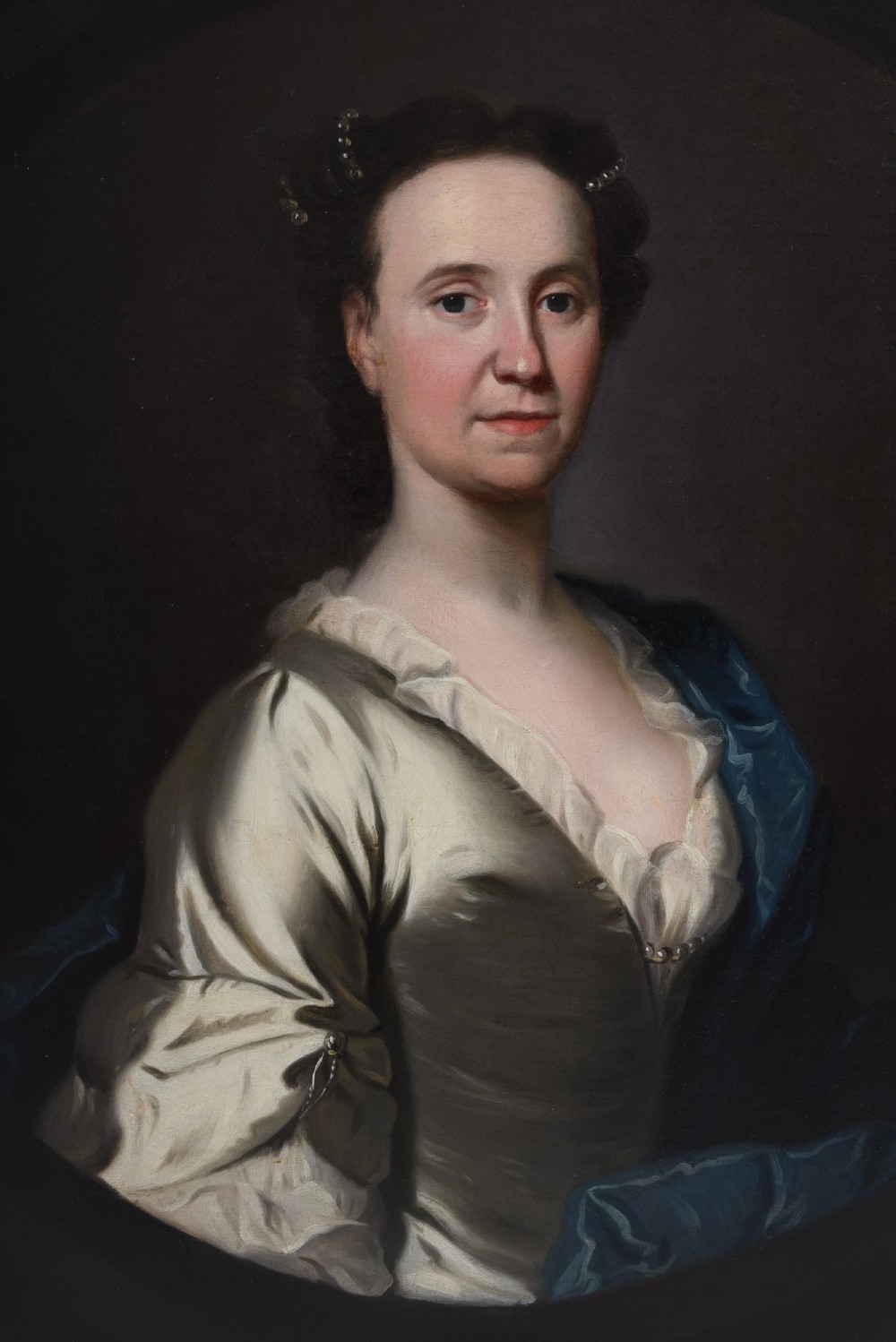 An 18th-century oil painting on canvas depicting a seated woman in a cream satin dress with a blue wrap, set within an ornate carved giltwood frame. View V