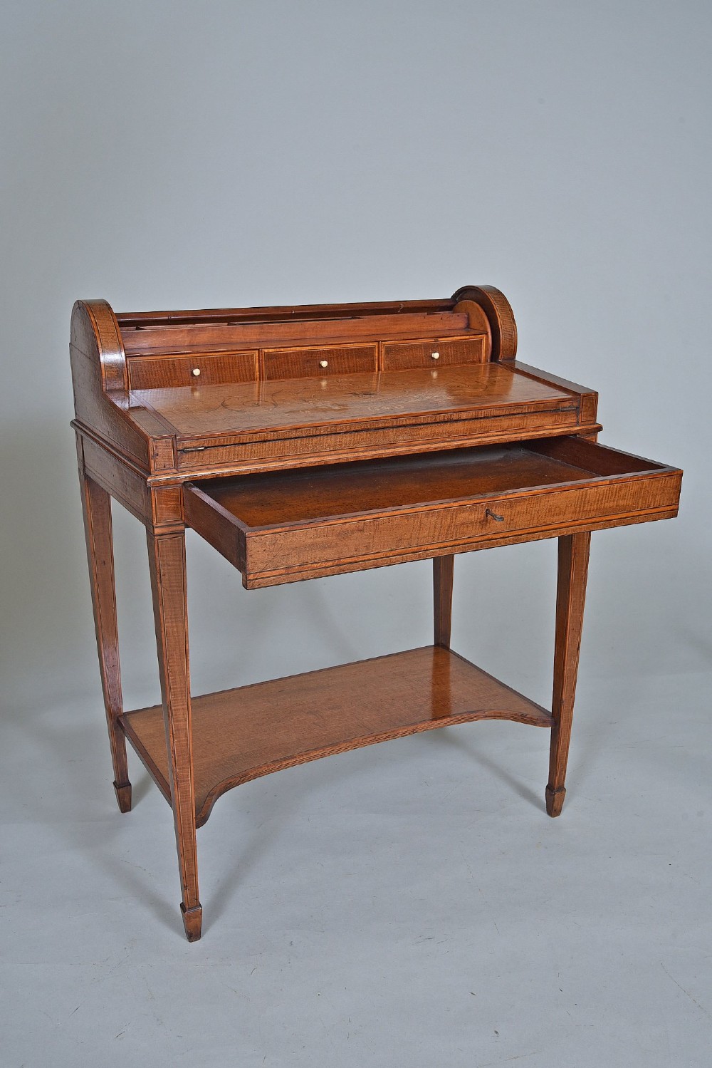 A 19th-century satinwood cylinder bureau featuring an inlaid curved roll-top hood, frieze drawer, square tapered legs, and a shaped lower shelf. View IV