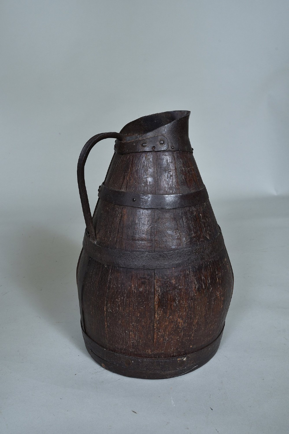 A dark brown 19th-century staved oak harvest jug featuring wrought iron bands, a pour spout, and a curved metal handle.