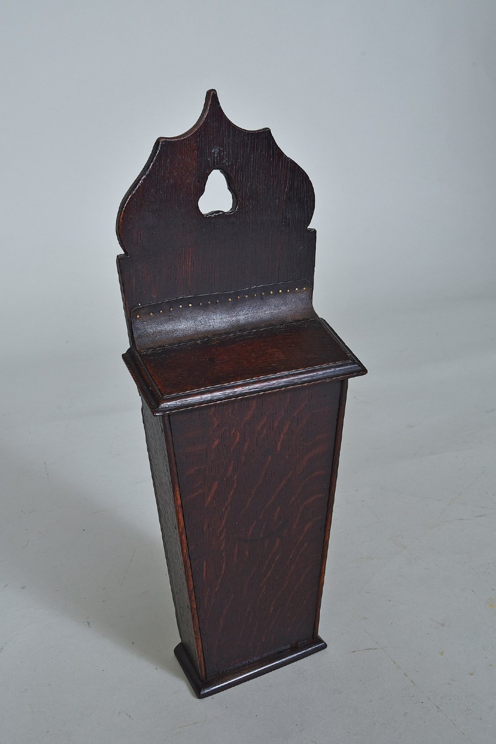 A dark oak wall-hanging candle box featuring a shaped backboard with a hanging aperture, a sliding front cover, and a tapered body.