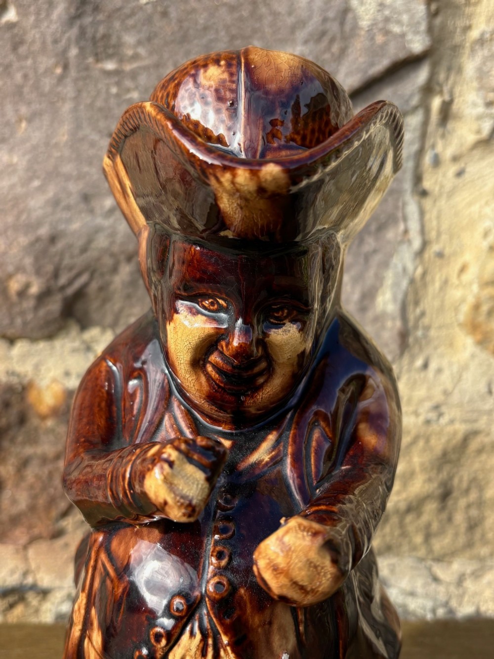 A nineteenth-century ceramic Toby jug depicting a seated man wearing a tricorne hat, finished in a rich, mottled treacle-brown glaze. View VI