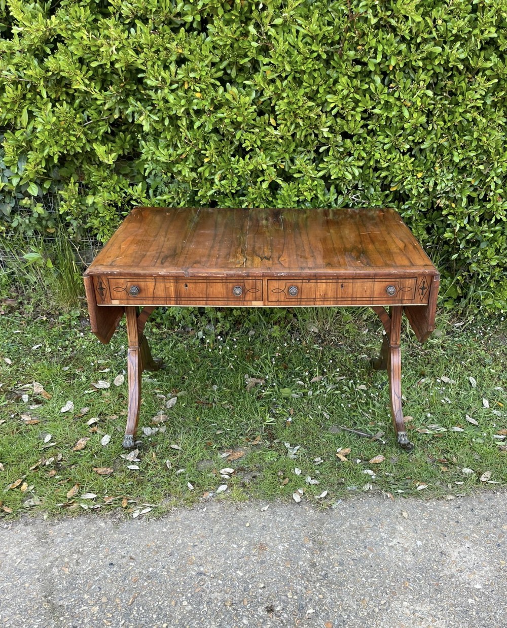 An antique Regency mahogany sofa table featuring drop-leaf side flaps, inlaid drawer fronts with brass handles, and splayed legs ending in brass paw castors.