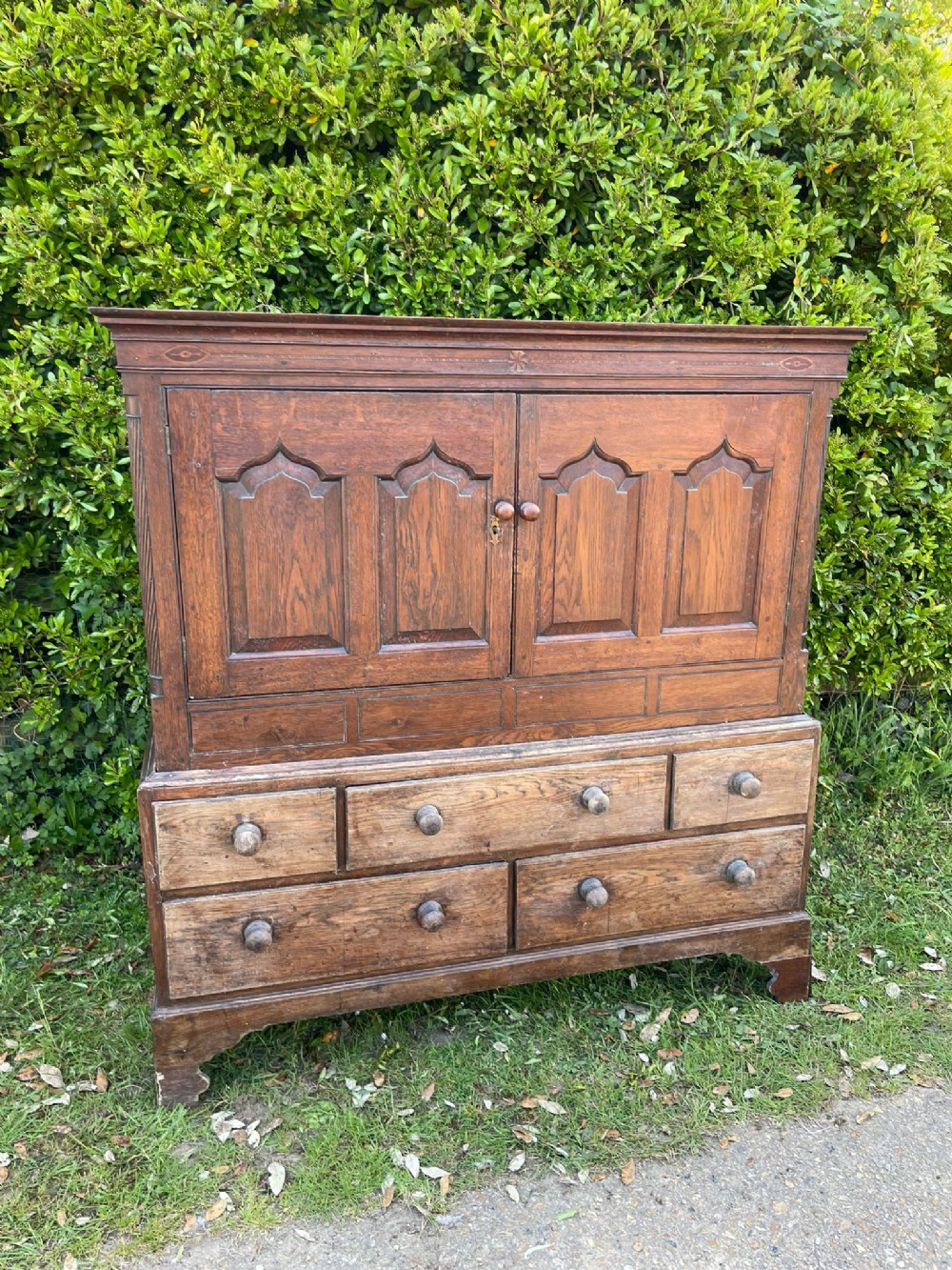 An 18th-century oak hall press cupboard featuring double doors with arched fielded panels, an inlaid frieze, and a base containing multiple drawers with turned wooden handles.
